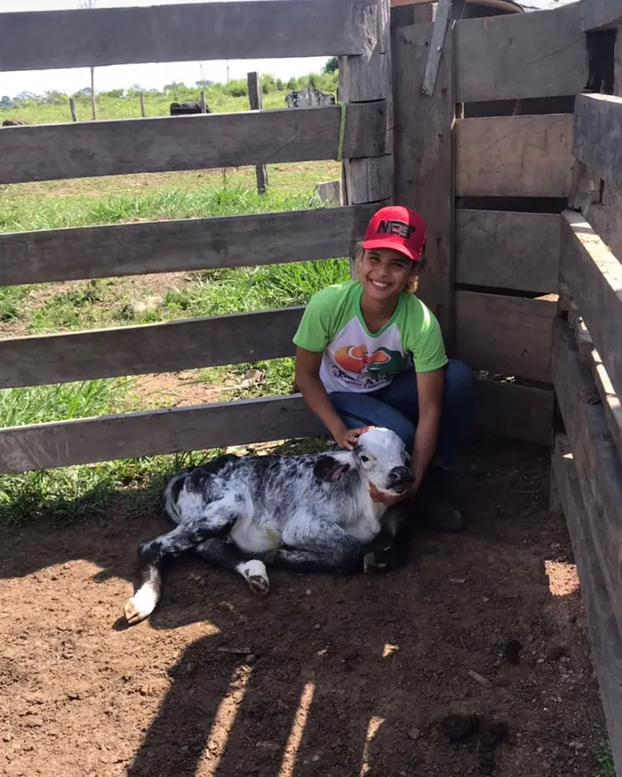 Tháscila agachada no curral, sorrindo, ao lado de um bezerro