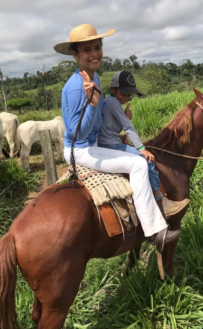 Tháscila a cavalo, de chapéu de palha, com uma criança na garupa, num pasto da vicinal